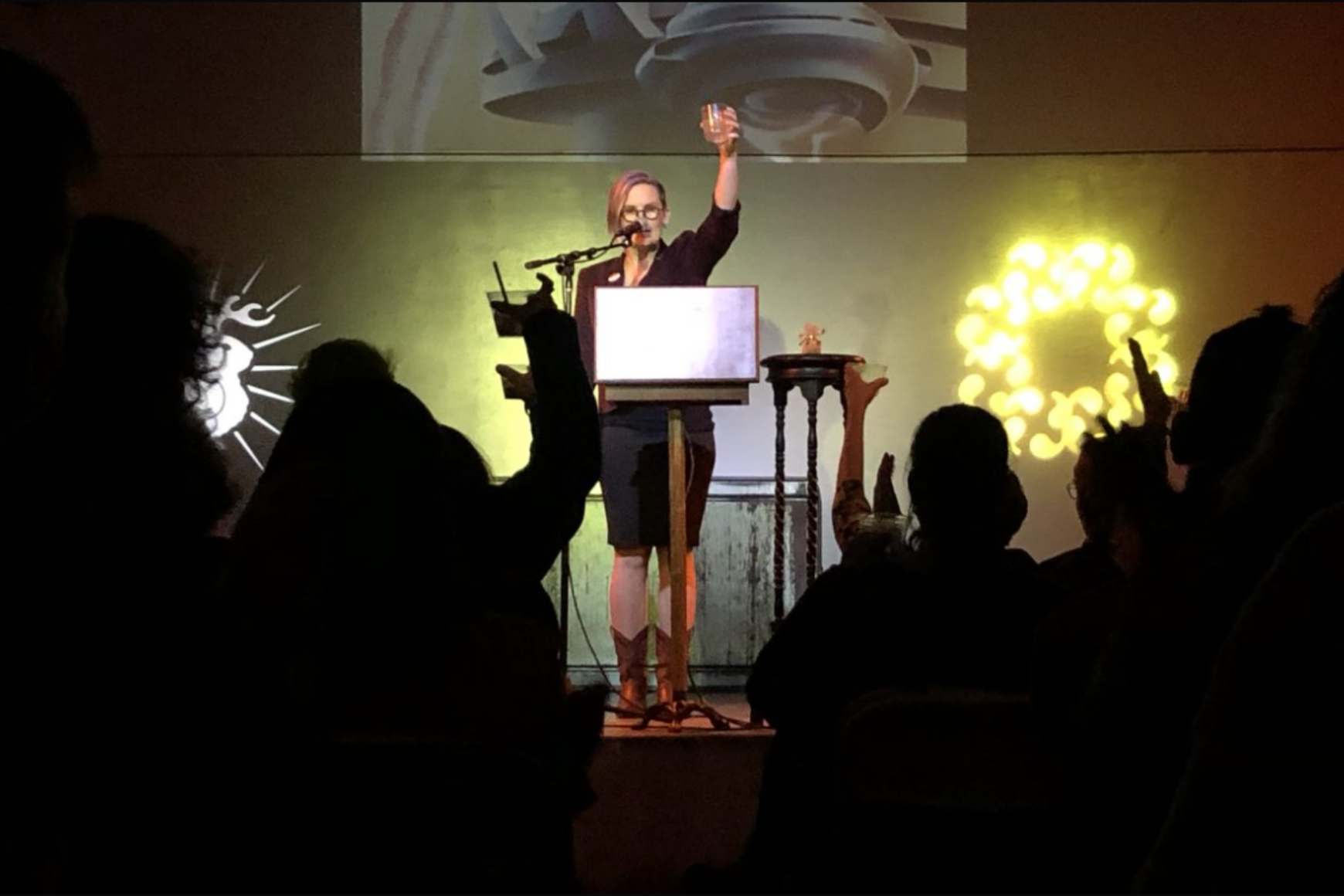 A speaker on stage raises a glass in a toast, while an audience in silhouette joins in, illuminated by warm stage lighting and projected visuals.